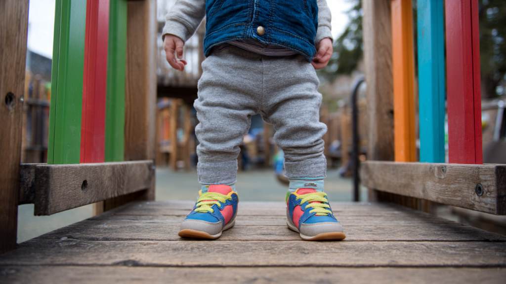 A toddler taking first steps in colorful wide toe box sneakers on a playground, smiling with confident balance.